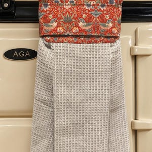 May include: A hanging kitchen towel featuring a waffle-textured light grey fabric attached to a top band with a vibrant red background and a repeating pattern of stylised birds, flowers, and foliage in shades of blue, cream, and muted green.