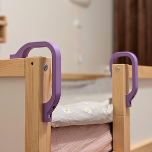 Puede incluir: Primer plano de una estructura de cama con postes de madera clara y paneles blancos. La estructura de la cama tiene asas de seguridad moradas. La ropa de cama incluye una sábana blanca con estampado floral y una manta rosa.