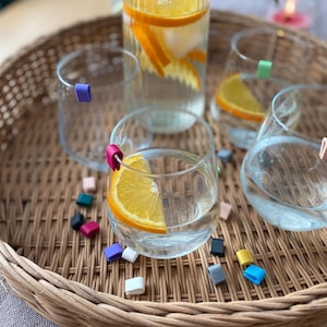 May include: A clear glass water bottle and glasses with orange slices, set on a woven tray. The glasses have colorful drink markers. A variety of small, colorful drink markers are scattered on the tray.