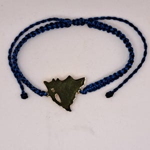 Puede incluir: Una pulsera de macramé azul oscuro con un dije dorado en forma de murciélago. La pulsera tiene nudos corredizos ajustables y está hecha de cordón azul oscuro. El dije tiene un diseño recortado.