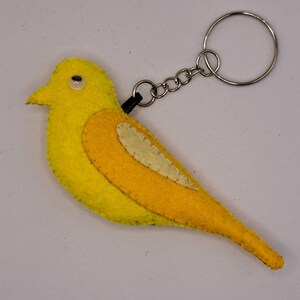 May include: A yellow felt bird keychain with a silver metal keyring and chain. The bird has a white and yellow design on its body, a black eye, and a black attachment to the chain. The keychain is a cheerful accessory.
