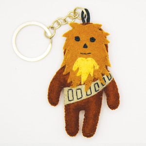 Könnte beinhalten: Ein brauner Filz-Chewbacca-Schlüsselanhänger mit einem gelben Filzbart und einem schwarzen Filz-Bandolier. Der Schlüsselanhänger hat einen goldenen Schlüsselring aus Metall.