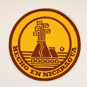 May include: A round, yellow embroidered patch with a brown volcano and tree design. The text "HECHO EN NICARAGUA" is written around the edge of the patch.