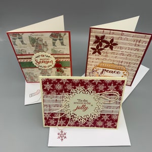 May include: Three handmade Christmas cards with decorative elements. One card features a Santa Claus design, another has musical notes and burgundy flowers, and the third has snowflakes and the text "'Tis the season to be jolly". Each card is in a white envelope.