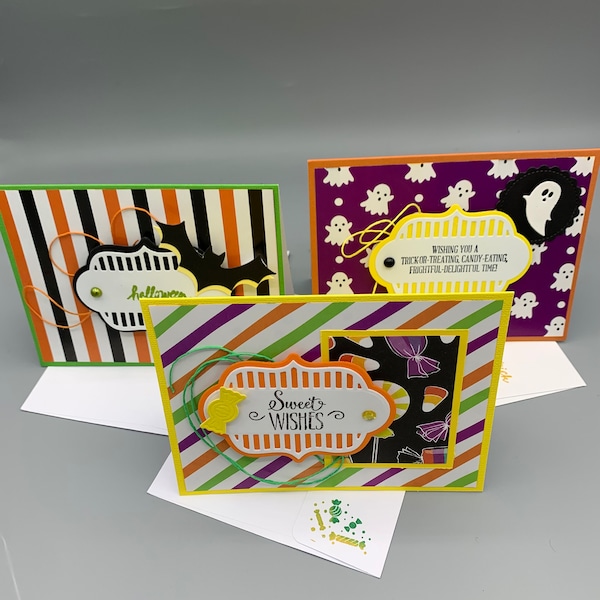 Puede incluir: Tres tarjetas de felicitación de Halloween con diferentes diseños. La primera tarjeta tiene rayas blancas y negras con un murciélago y la palabra "halloween" en ella. La segunda tarjeta tiene un fondo rayado morado y verde con un marco amarillo y naranja que dice "Sweet Wishes". La tercera tarjeta tiene un fondo morado con fantasmas blancos y una etiqueta amarilla que dice "Wishing you a trick-or-treating candy-eating frightful-delightful time!"