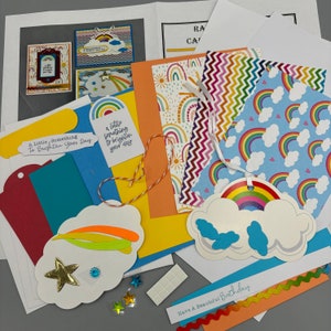 May include: A collection of rainbow-themed craft supplies for card making. The supplies include patterned paper, cardstock, die cuts, and embellishments. The text "A Little Something To Brighten Your Day" and "Have A Beautiful Birthday" are included.