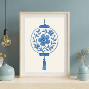 May include: A framed watercolor print featuring a blue floral lantern design. The lantern is round with a central flower and surrounding floral and leaf accents. A tassel hangs from the bottom. The artwork is set against a cream background.