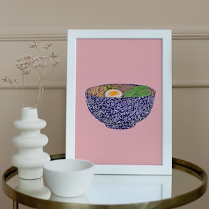 May include: A framed print of a blue and white bowl of ramen with an egg, green onions, and other toppings on a pink background.