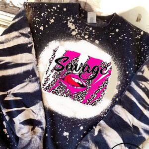 May include: Black sweatshirt with a white and pink graphic design that says "Savage" with a pink tongue sticking out of a pink mouth. The sweatshirt has a bleach splattered design.