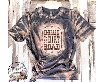 May include: A dark grey t-shirt with a bleach-splattered design. The shirt features a brown graphic with barbed wire around the edges that says "Chillin' on a Dirt Road".