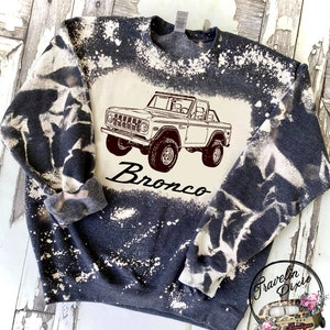 May include: A dark blue sweatshirt with a white bleach design. The design features a vintage Ford Bronco with the word "Bronco" underneath.