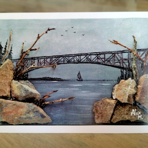 May include: A gray and blue painting of a bridge over water with a sailboat in the distance. The painting is decorated with small branches and rocks.