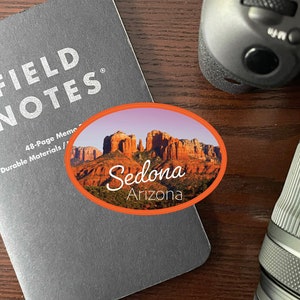May include: A gray Field Notes memo book with the text "FIELD NOTES" and an oval sticker featuring a Sedona, Arizona landscape. A camera lens and camera body are also visible on a wooden surface.