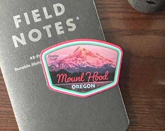 May include: A black Field Notes notebook with the text "FIELD NOTES" and a colorful sticker featuring Mount Hood, Oregon. A camera lens and camera body are also visible.
