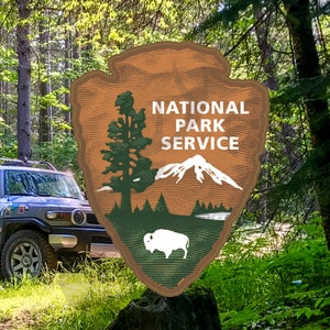 May include: A gray SUV with a world map sticker design parked in a forest. The vehicle has a roof rack and a National Park Service sticker on the back. The sticker is brown with a green pine tree, a white mountain, and a brown bison.