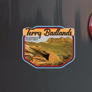 May include: A rectangular sticker with the text "Terry Badlands Wilderness Study Area" and an image of a landscape with rock formations and green grass. Below is a sticker with the text "Stuck On Travel VINYL DECALS TRAVEL MEMORIES".