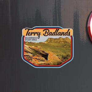 May include: A rectangular sticker with the text "Terry Badlands Wilderness Study Area" and an image of a landscape with rock formations and green grass. The sticker is on a dark gray surface. Below is a sticker that says "Stuck On Travel Vinyl Decals Travel Memories".