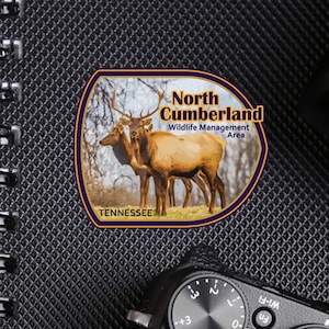 May include: A brown and gold vinyl decal sticker with a picture of two elk in a forest setting. The sticker says "North Cumberland Wildlife Management Area" and "Tennessee". The sticker is on a black notebook with a camera on top of it.