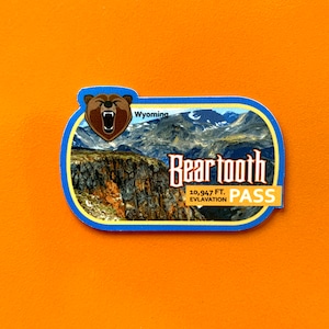 May include: A blue and white sticker with a cartoon bear and the text "Beartooth Pass" with the elevation of 10,947 feet. The sticker also includes a scenic mountain landscape and the word "Wyoming".
