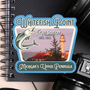 May include: A blue and white sticker with the text "Whitefish Point Light Station Since 1861" and "Michigan's Upper Peninsula". The sticker features a cartoon illustration of a fish and a lighthouse.