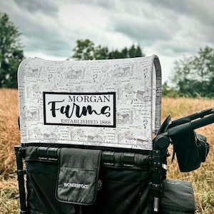 May include: A black and gray WonderFold wagon with a canopy featuring a farm-themed pattern. The canopy has the words "Morgan Farms Established 1888" in a black rectangle. The wagon has black wheels and storage pockets.
