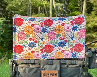 May include: A black wagon with a floral patterned canopy. The wagon has two large wheels and a black handle. The canopy is made of fabric and has a white background with colourful flowers. The wagon has a brown leather patch with the text "NORTH WOODS crafting co."
