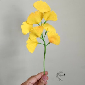 May include: A decorative stem featuring multiple bright yellow ginkgo biloba leaves attached to a thin green branch. The fan-shaped leaves have a textured appearance and are arranged along the length of the stem, which is held upright.