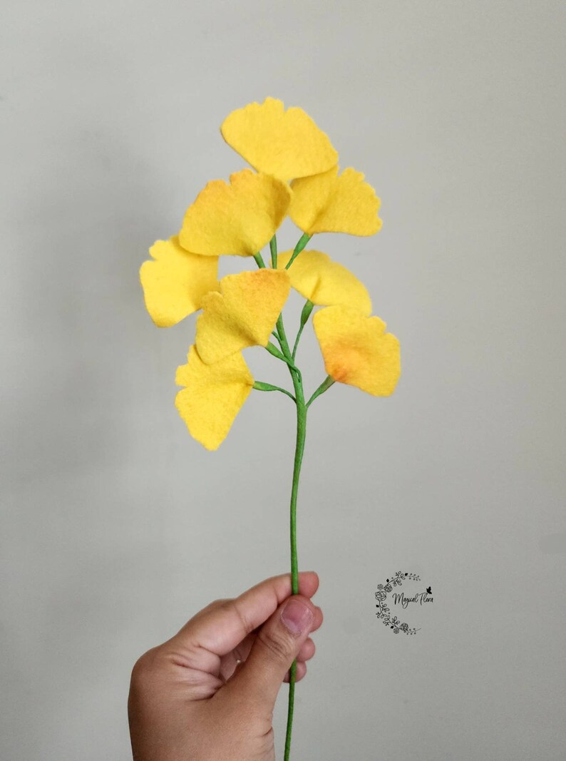 May include: A decorative stem featuring multiple bright yellow ginkgo biloba leaves attached to a thin green branch. The fan-shaped leaves have a textured appearance and are arranged along the length of the stem, which is held upright.