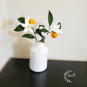 May include: A white ceramic vase containing a decorative branch with felt orange fruit, dark green leaves, and white blossoms with yellow centers. The branch features several small white buds and two round orange fruits attached to stems.