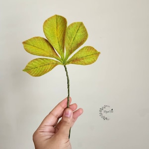 May include: A handcrafted five-lobed leaf sculpture featuring textured lime green and yellow fabric with reddish-brown edges, attached to a thin, wrapped green wire stem. A small circular logo with the text Magical Flora is visible.