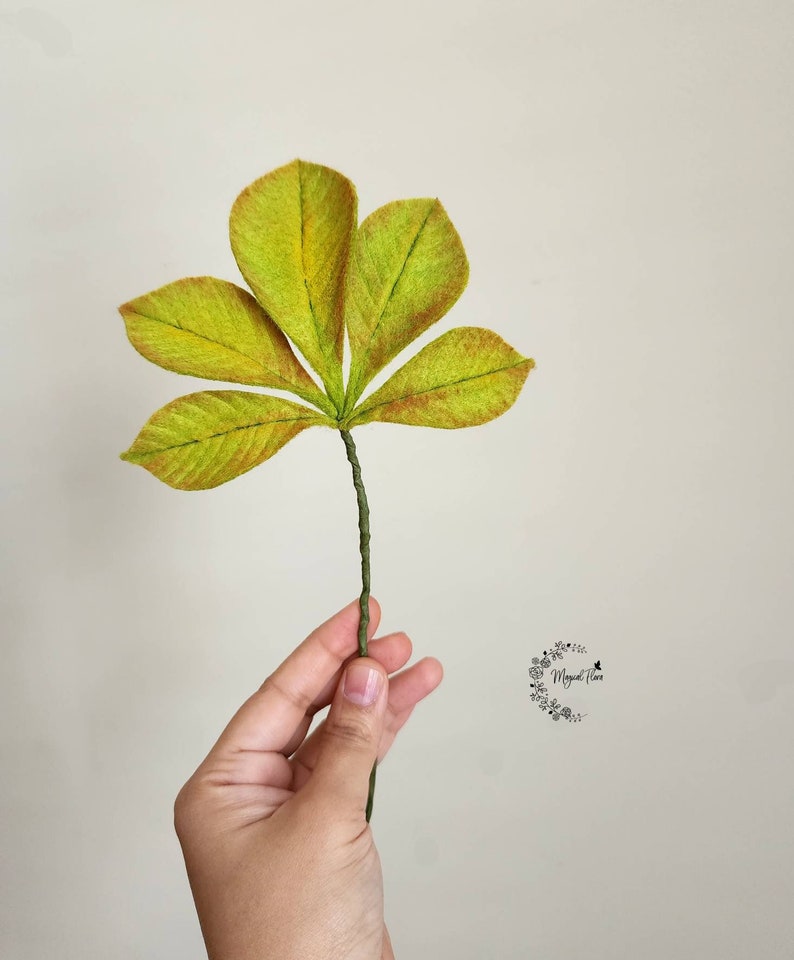 May include: A handcrafted five-lobed leaf sculpture featuring textured lime green and yellow fabric with reddish-brown edges, attached to a thin, wrapped green wire stem. A small circular logo with the text Magical Flora is visible.