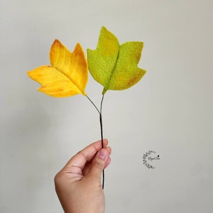 May include: Two felt leaves attached to a thin brown stem. One leaf is bright yellow with orange shading, and the other is light green with yellow and brown accents. A small circular logo with the text "Magical Flora" is visible on the right.