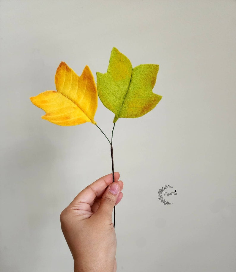 May include: Two felt leaves attached to a thin brown stem. One leaf is bright yellow with orange shading, and the other is light green with yellow and brown accents. A small circular logo with the text "Magical Flora" is visible on the right.