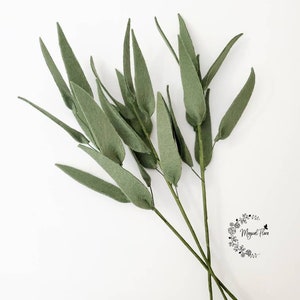 May include: Two stems of artificial eucalyptus foliage featuring elongated, pointed sage green leaves attached to thin, dark green wire branches. A small circular logo with the text Magical Flora is visible in the lower right corner.