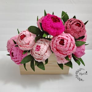 May include: A bouquet of felt flowers in shades of pink, ranging from pale blush to vibrant magenta, with dark green felt leaves. The flowers are arranged in a light-colored wooden rectangular box. A logo with the text Magical Flora is visible.