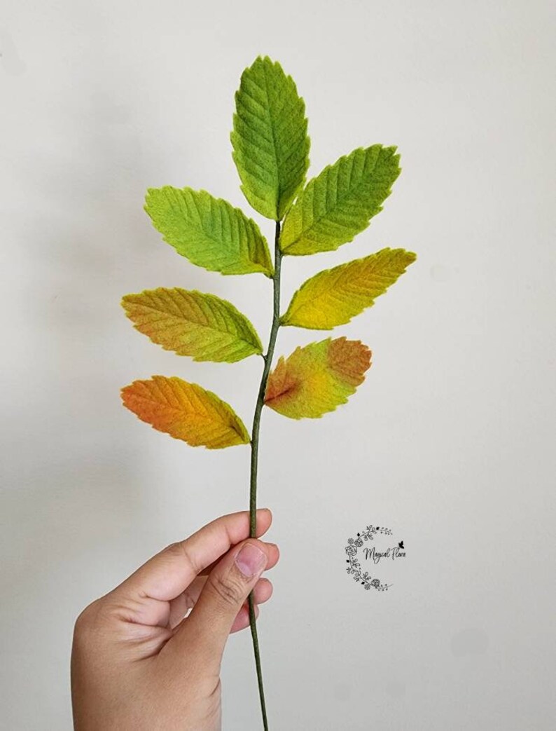 May include: A decorative botanical stem featuring seven serrated leaves with a gradient color transition from vibrant green at the top to yellow and orange at the base. The leaves have a textured surface and are attached to a thin, dark green stem.