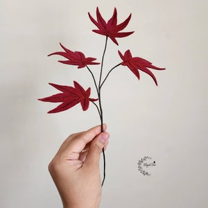 May include: A decorative branch featuring four deep red felt maple leaves with visible stitched vein details. The leaves are attached to a thin, dark brown stem. A small circular logo with floral elements and the text "Magical Flora" is visible.