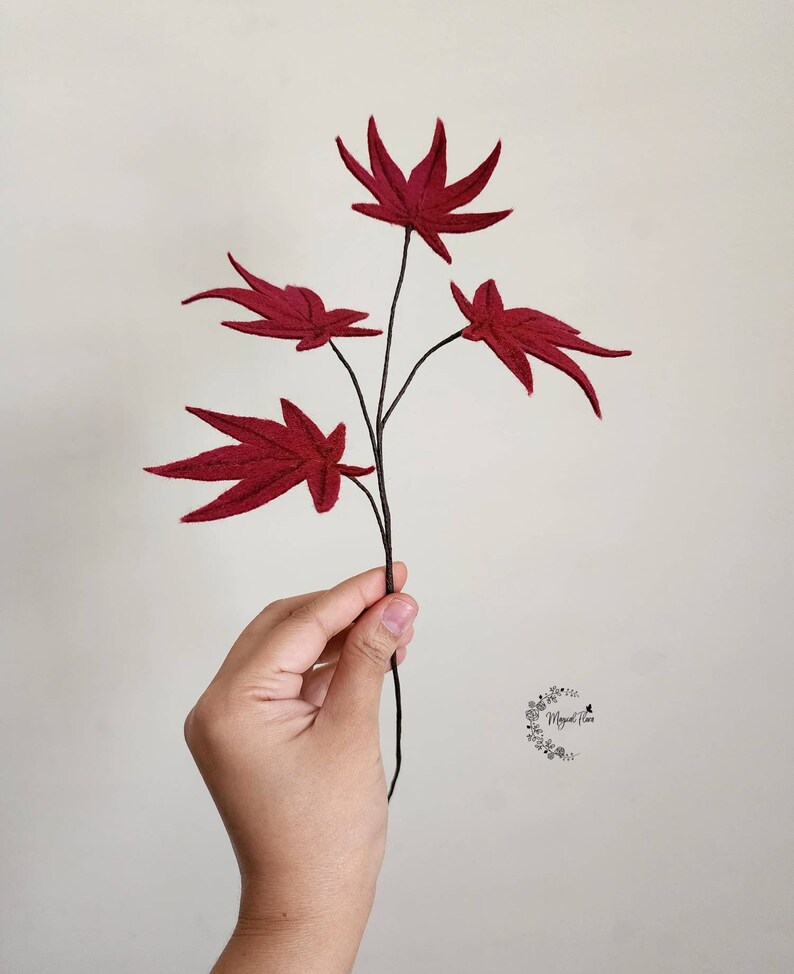 May include: A decorative branch featuring four deep red felt maple leaves with visible stitched vein details. The leaves are attached to a thin, dark brown stem. A small circular logo with floral elements and the text "Magical Flora" is visible.