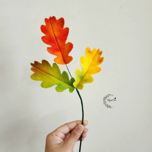 May include: A decorative stem featuring three felt oak leaves in vibrant autumn colors. The top leaf is orange, the left leaf is green and brown, and the right leaf is yellow and green. A small circular logo with floral details is visible on the right.