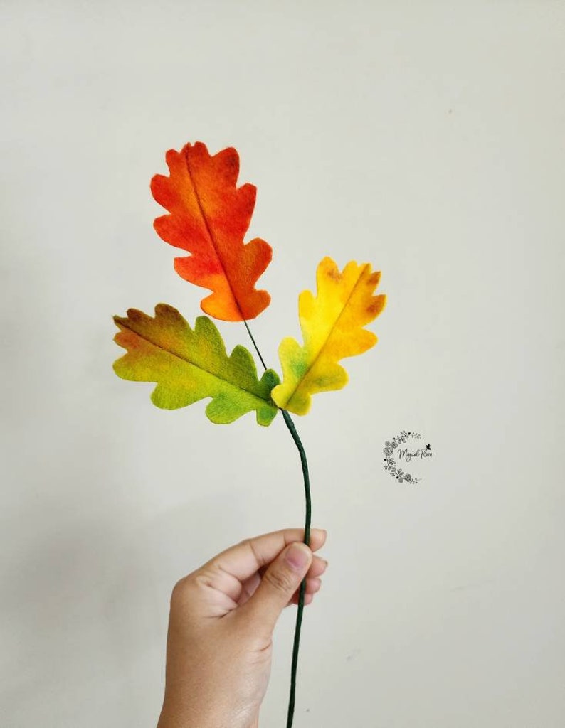 May include: A decorative stem featuring three felt oak leaves in vibrant autumn colors. The top leaf is orange, the left leaf is green and brown, and the right leaf is yellow and green. A small circular logo with floral details is visible on the right.