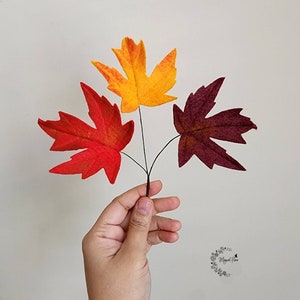 May include: Three artificial maple leaves attached to thin dark stems, featuring one bright red leaf, one vibrant yellow-orange leaf, and one deep burgundy leaf. The leaves are arranged in a fan shape, displaying distinct pointed lobes and veins.