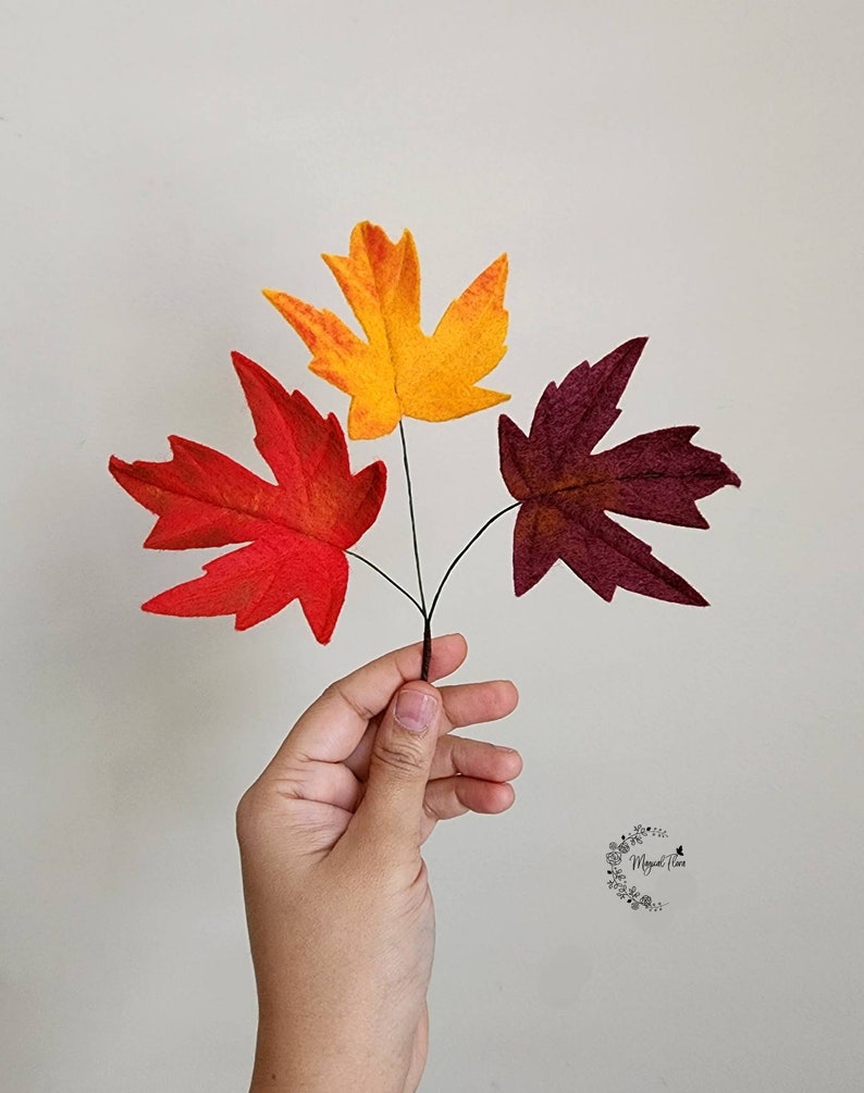 May include: Three artificial maple leaves attached to thin dark stems, featuring one bright red leaf, one vibrant yellow-orange leaf, and one deep burgundy leaf. The leaves are arranged in a fan shape, displaying distinct pointed lobes and veins.