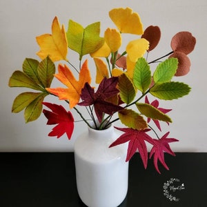 May include: A white ceramic vase containing a bouquet of felt leaves in various autumn colors including yellow, orange, red, brown, and green. The leaves are attached to thin wire stems, creating a realistic arrangement of foliage in shades of fall.