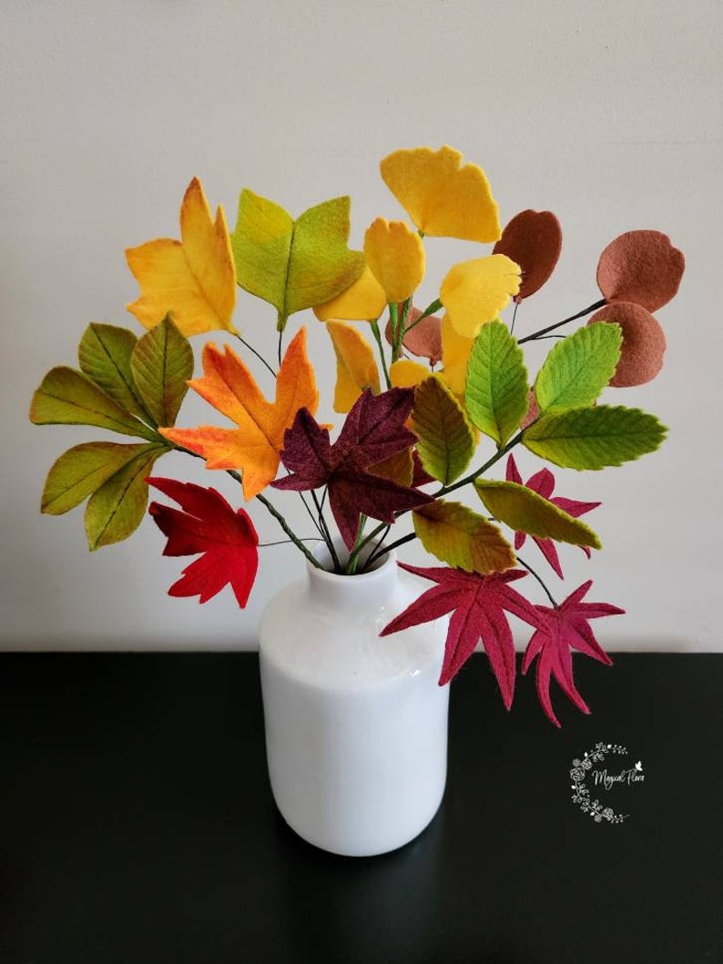 May include: A white ceramic vase containing a bouquet of felt leaves in various autumn colors including yellow, orange, red, brown, and green. The leaves are attached to thin wire stems, creating a realistic arrangement of foliage in shades of fall.