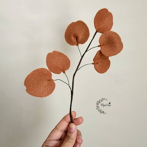 May include: A decorative branch featuring six round, rust-colored felt leaves attached to a thin, dark brown stem. The leaves have a soft, textured appearance and are arranged symmetrically along the branch. A small logo is visible on the right.
