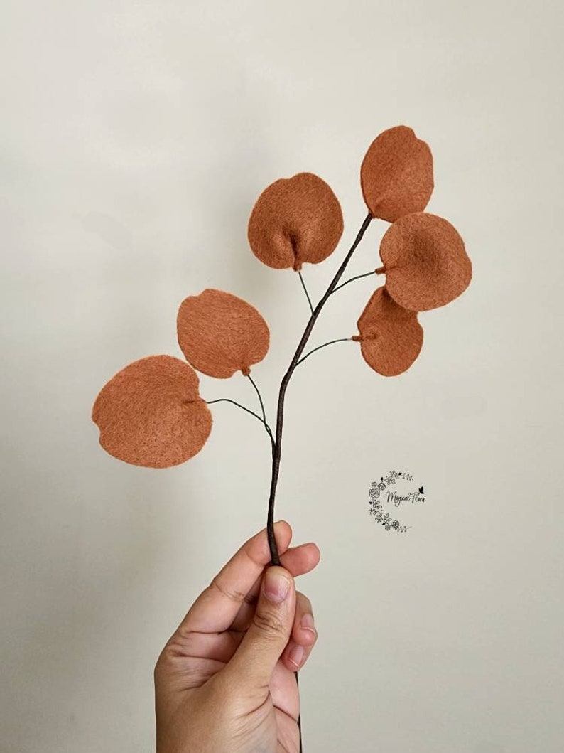 May include: A decorative branch featuring six round, rust-colored felt leaves attached to a thin, dark brown stem. The leaves have a soft, textured appearance and are arranged symmetrically along the branch. A small logo is visible on the right.