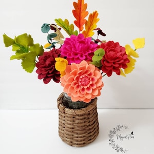 May include: A bouquet of felt flowers in shades of hot pink, peach, and deep red, accented with green and yellow felt leaves and orange oak-shaped foliage. The flowers are arranged in a brown woven basket. The text Magical Flora is visible.