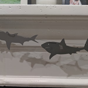 May include: A string of paper shark cutouts, including a hammerhead shark and a great white shark, hanging from a white ceiling.