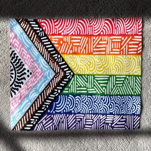 May include: A vibrant textile displaying a Pride flag design. The flag includes the classic rainbow colours, plus stripes representing the transgender community and people of colour. The design is made up of diverse geometric patterns.