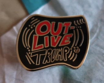 May include: A black and gold enamel pin with the words "OUT LIVE TRUMP" in red and white lettering. The pin has a unique shape with decorative lines and dots.